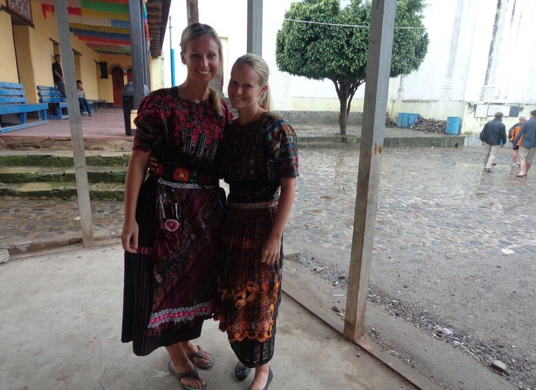 Two women standing in a covered outdoor area with a cobblestone ground, a tree, and a building with blue benches in the background. 2