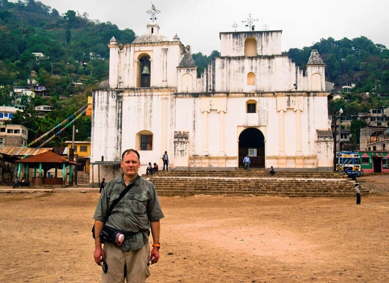 A man standing in front of a white colonial-style church with a bell tower, surrounded by a hilly landscape and residential buildings. 4