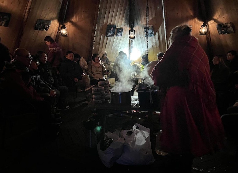 A group of people gathered around a central cooking area with steam rising, in a rustic indoor setting with wooden walls and hanging lanterns. 3