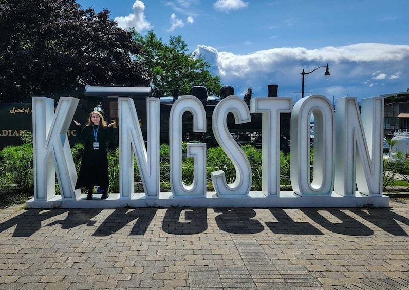 A large white sign with the word 'KINGSTON' in capital letters, a person standing in front of it, and a clear blue sky with clouds in the background. 5