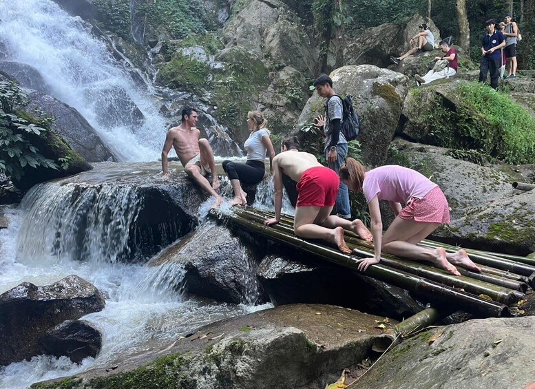People sitting on a log over a waterfall in a forested area. 2