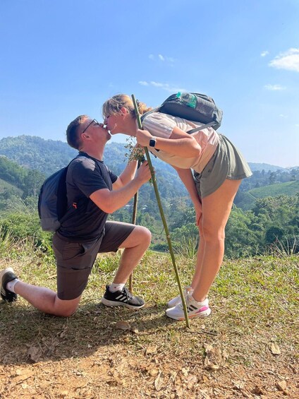 A person kneeling and holding a camera on a tripod, with another person standing beside them on a grassy hill with a forested background. 1