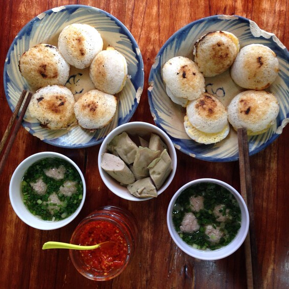 A wooden table with two bowls of white rice cakes, two bowls of soup with meatballs and green onions, and a small bowl of red sauce. 1