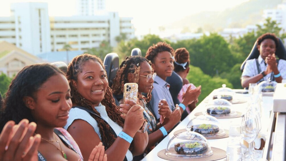 A group of people seated at an outdoor event with food and drinks on a long table. 2