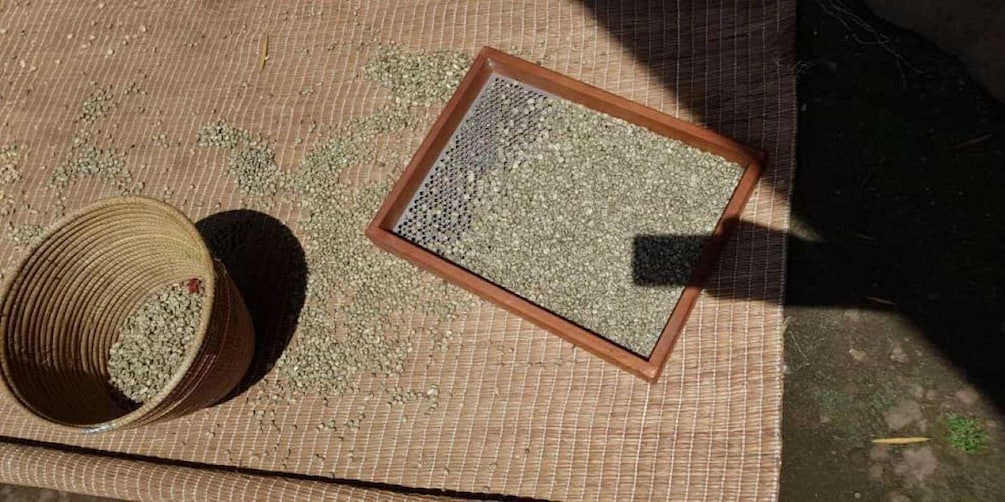 A woven mat with a rectangular container filled with small pebbles, a cylindrical object with a red marking, and a shadow cast on the mat. 2