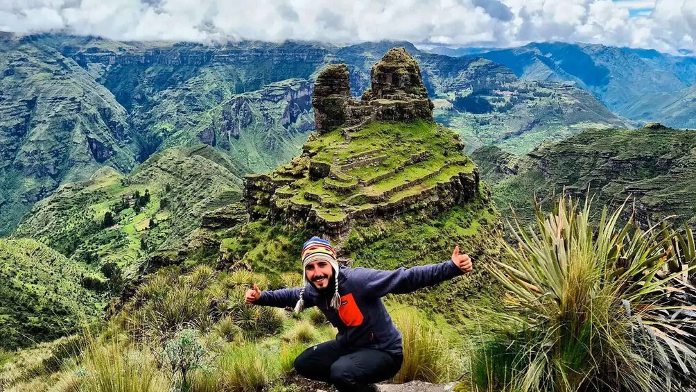 A person in a blue jacket with a red pocket, squatting on a grassy hill with a mountainous landscape in the background. 5