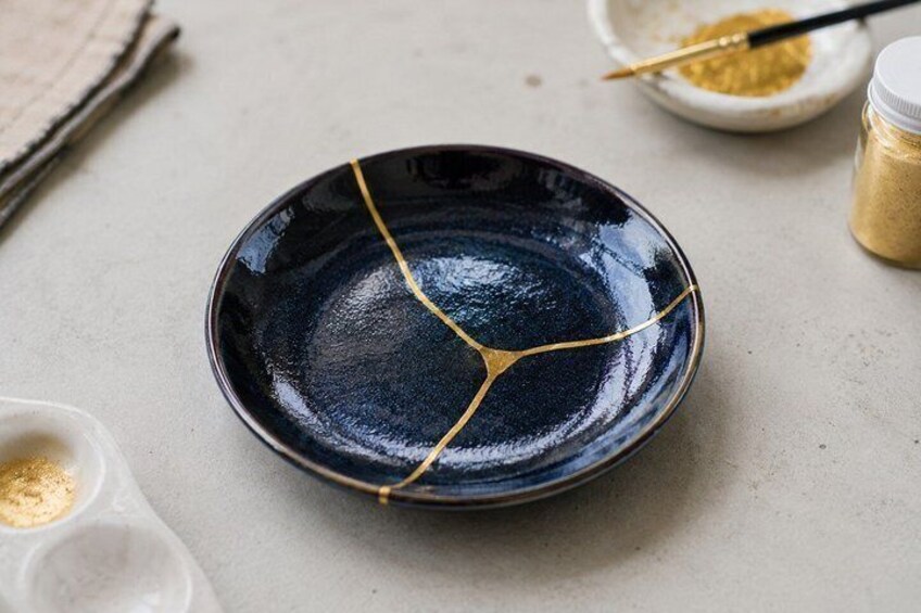 A dark blue ceramic plate with gold lines, a small bowl of yellow powder, and a paintbrush on a light surface. 4