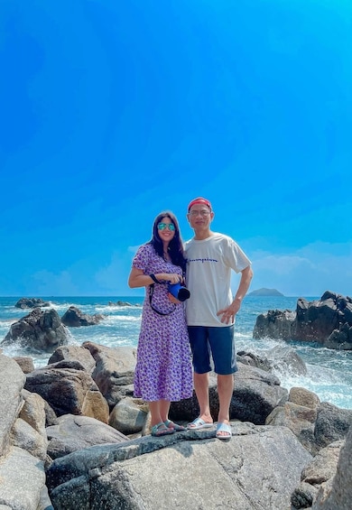 Two people standing on rocky terrain by the ocean with waves crashing against the rocks. 5