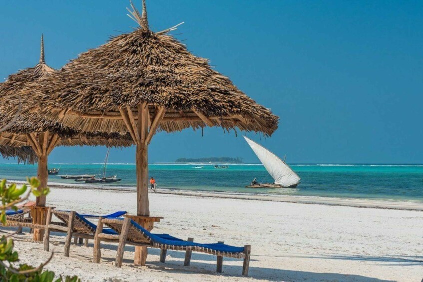 A beach with thatched umbrellas, lounge chairs, and a sailboat in the water. 3
