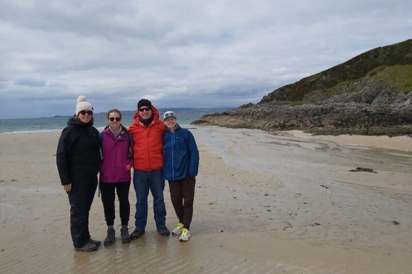 A group of people standing on a sandy beach with a rocky shoreline and overcast sky in the background. 4