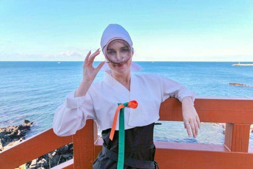 A person wearing a white outfit with a green and orange ribbon, standing by a wooden railing with a view of the sea and a distant island. 5