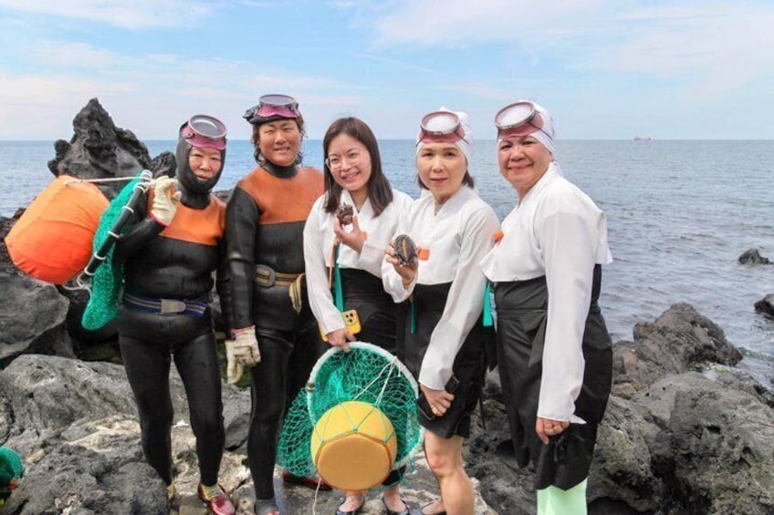 A group of people standing on rocky terrain by the sea, some wearing diving gear and holding diving equipment. 1