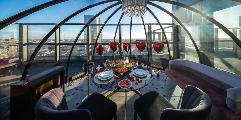 A high-ceilinged dining area with a table set for two, surrounded by heart-shaped balloons and a city view. 5
