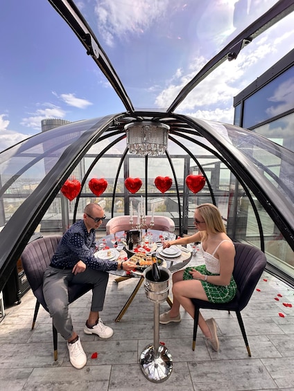 A couple dining inside a glass dome with a cityscape view, a chandelier, and heart-shaped decorations. 1