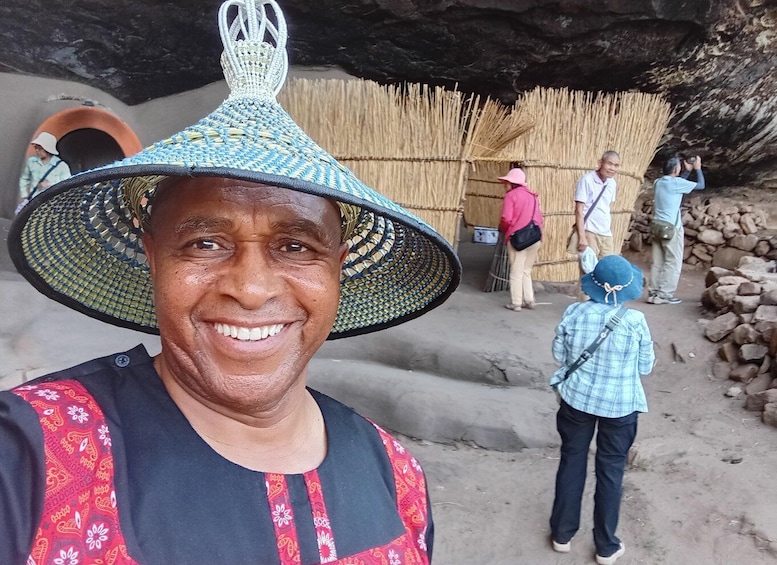 A person wearing a patterned hat and a dark shirt with red embroidery, standing in front of a cave entrance with a straw wall and a pile of rocks. 5