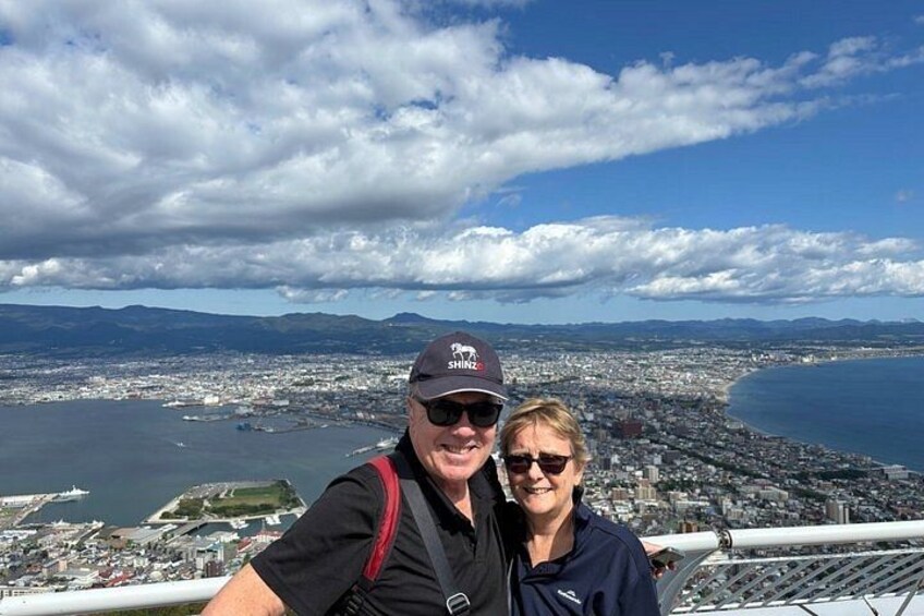 A person wearing a cap with 'SH' on it, standing on a high vantage point overlooking a coastal cityscape with mountains in the distance. 5