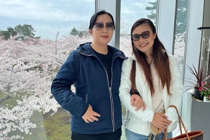 Two people standing indoors with a view of cherry blossoms outside. 2