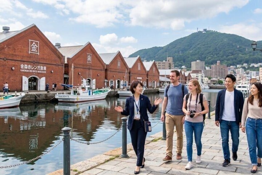 A group of people walking by a waterfront with brick buildings and a hill in the background. 1