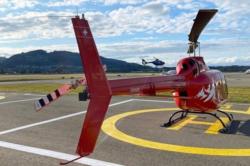 A red helicopter with a Swiss flag on a tarmac with yellow lines, another helicopter in the background, and mountains in the distance. 4