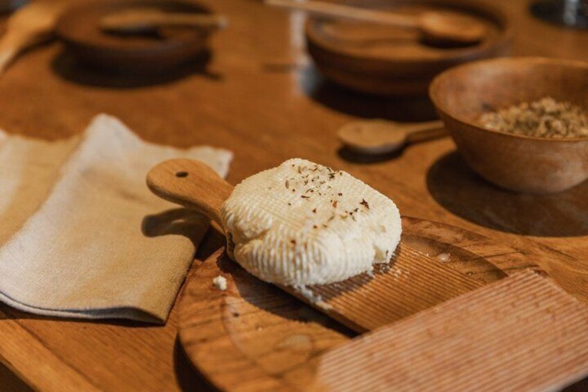 A cheese wheel on a wooden cutting board with a wooden knife and a bowl of grains in the background. 3