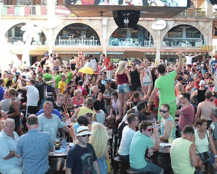 A crowded outdoor event with people seated at tables, a stage with a large screen, and a building with arches in the background. 1