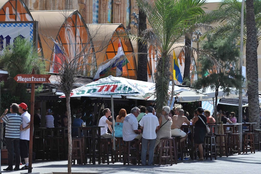 A crowded outdoor café with people dining and socializing, palm trees, and a colorful building in the background. 5