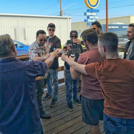 A group of people toasting with drinks on a wooden deck in an industrial area with shipping containers and a sign for 'Nomadic Beerworks.' 4