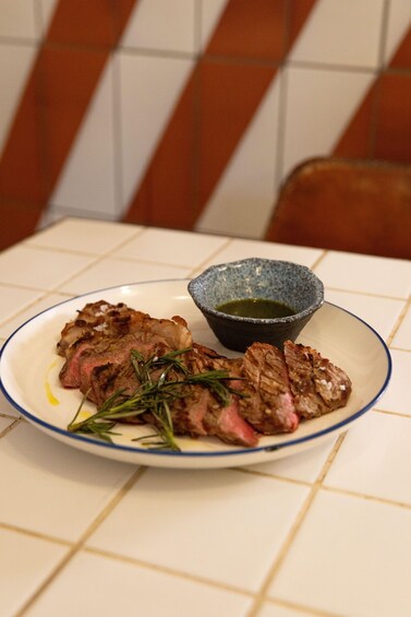 A plate of steak with rosemary and a small bowl of sauce on a tiled table. 4