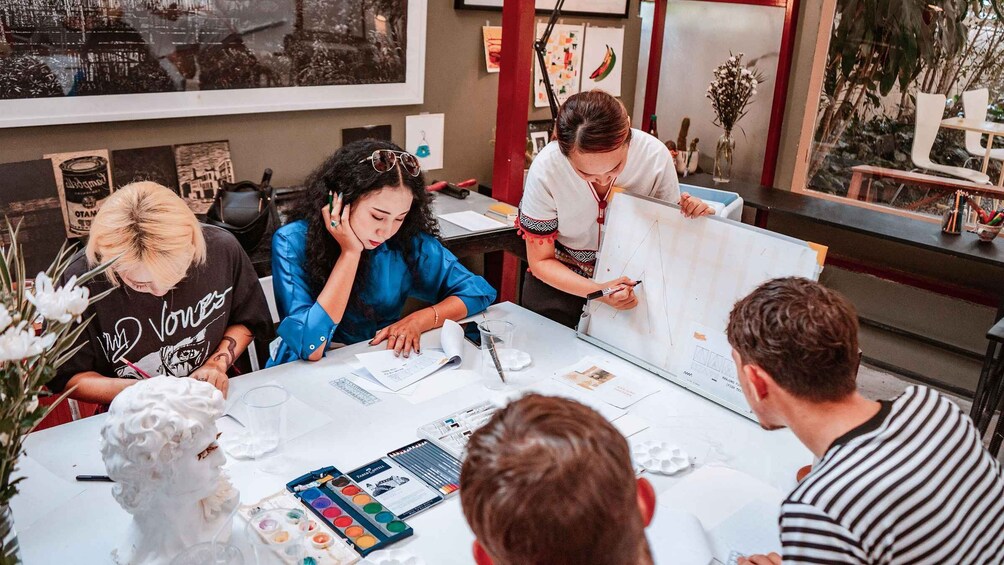 A group of people engaged in a collaborative design session around a large whiteboard, with art supplies and reference materials on the table. 2