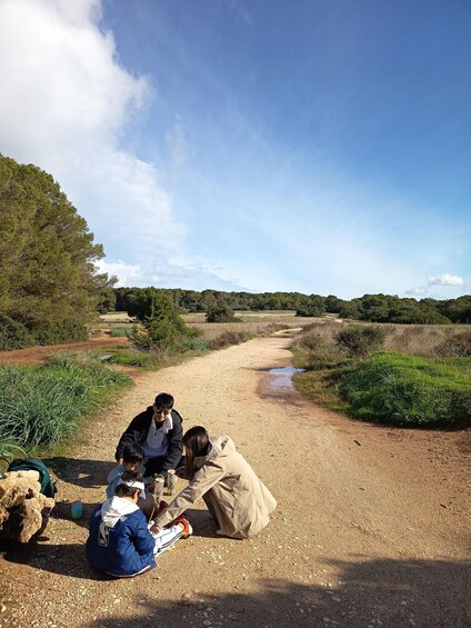 A group of people sitting on a gravel path in a natural landscape with trees and a clear sky. 5
