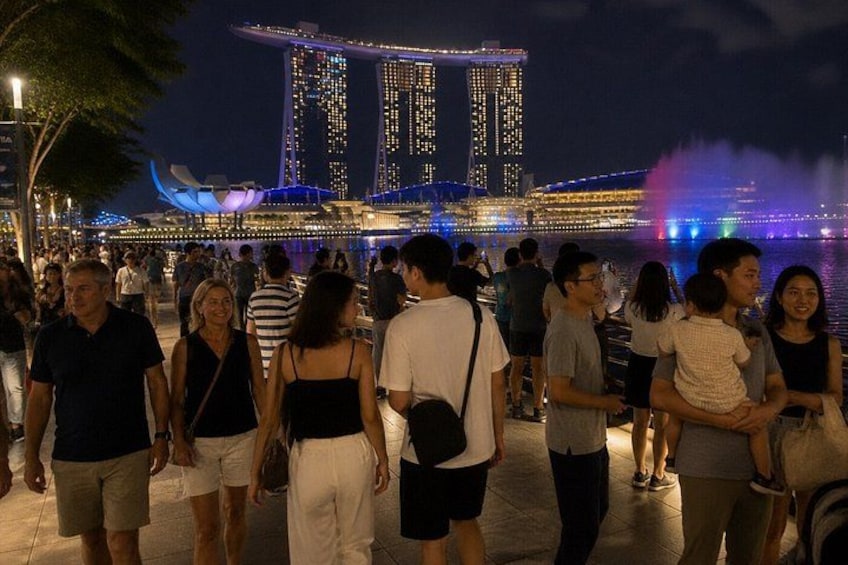 A crowded waterfront with people walking, a prominent illuminated building, and a colorful fountain display. 2