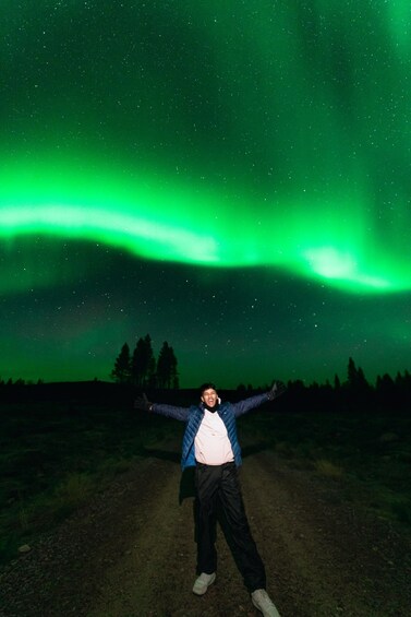 Une personne se tient debout sur un chemin de terre, les bras tendus, sous un ciel nocturne illuminé d’une aurore boréale verte. 3