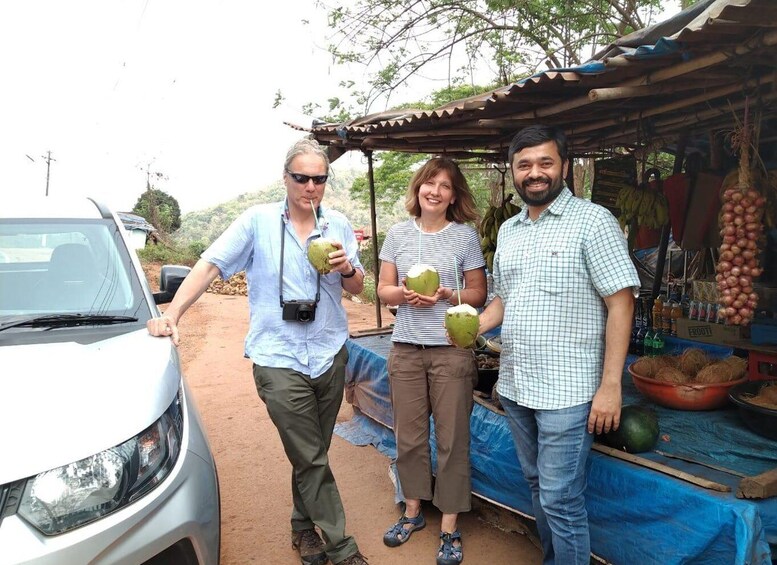 Three people holding coconuts at a roadside stall with a car parked nearby. 2