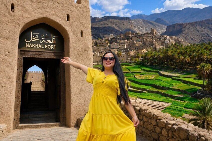 A woman in a yellow dress standing in front of Nakhhal Fort with a view of the village and mountains in the background. 1