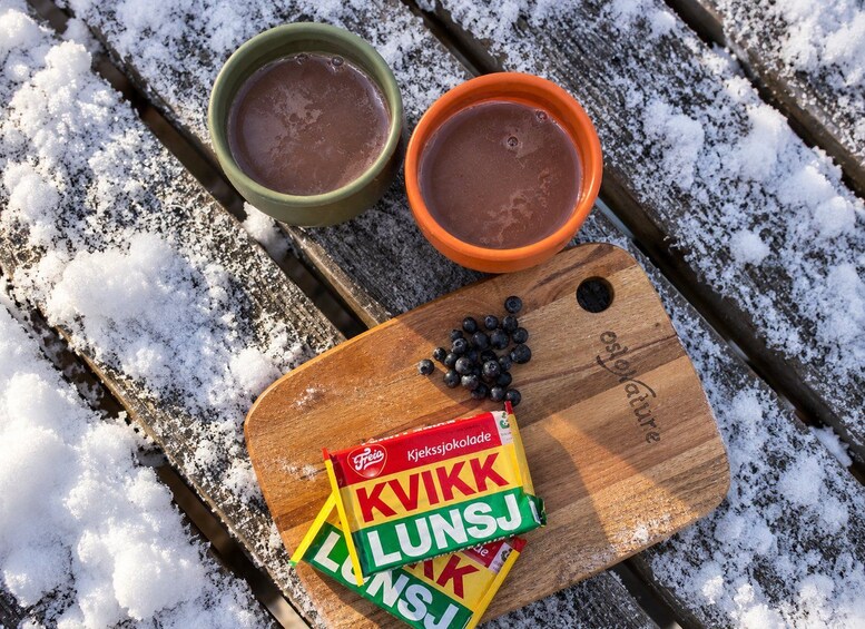 Two cups of hot chocolate on a wooden bench with snow around. There's a packet of Kveik chocolate and some blueberries on the bench. 5