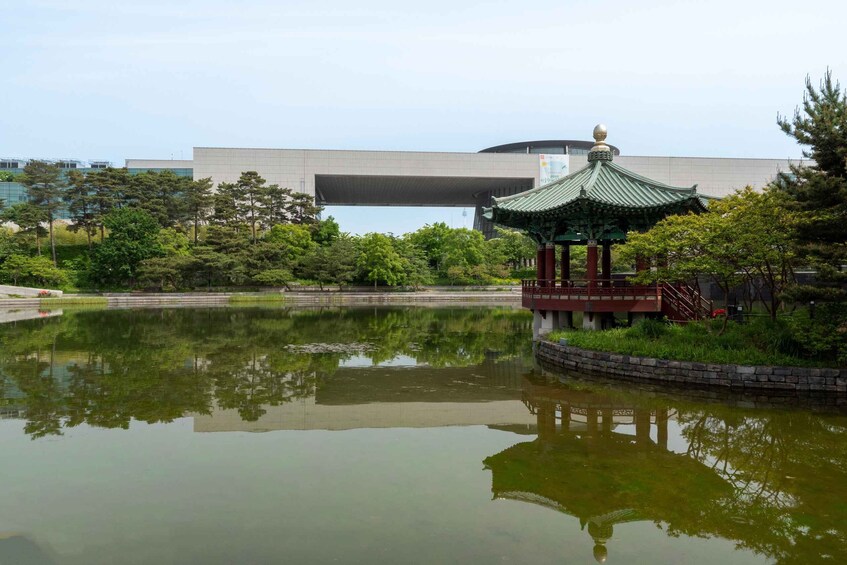 Un étang paisible avec un pavillon de style traditionnel, entouré d’une végétation luxuriante et, au loin, un bâtiment moderne. 4