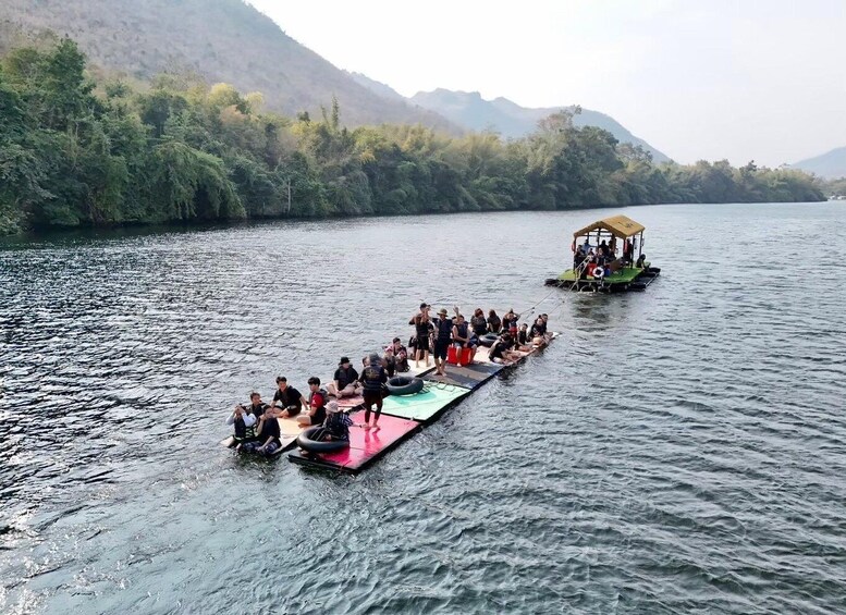A group of people on a long raft floating on a river, with a smaller boat in the background. 3