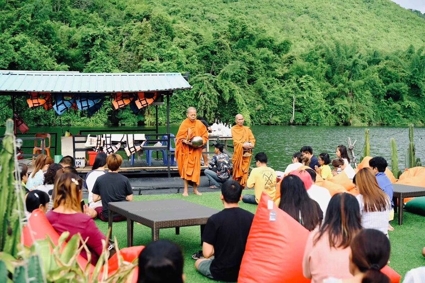 A group of people seated outdoors, some in orange robes, near a body of water with a forested hill in the background. 5