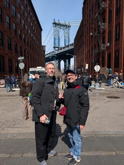 Two people standing in a city square with a bridge in the background. 2