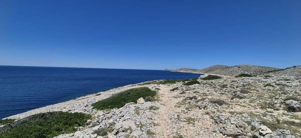 A coastal landscape with a clear blue sea, rocky terrain, and sparse vegetation. 4