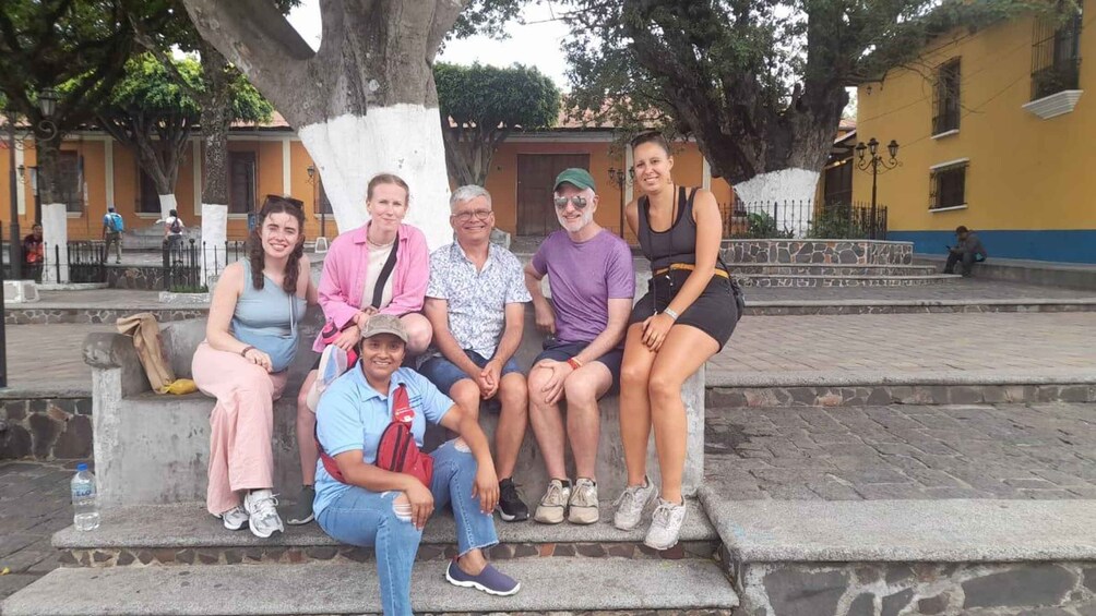 A group of people sitting on stone steps in a public square with trees and buildings in the background. 2