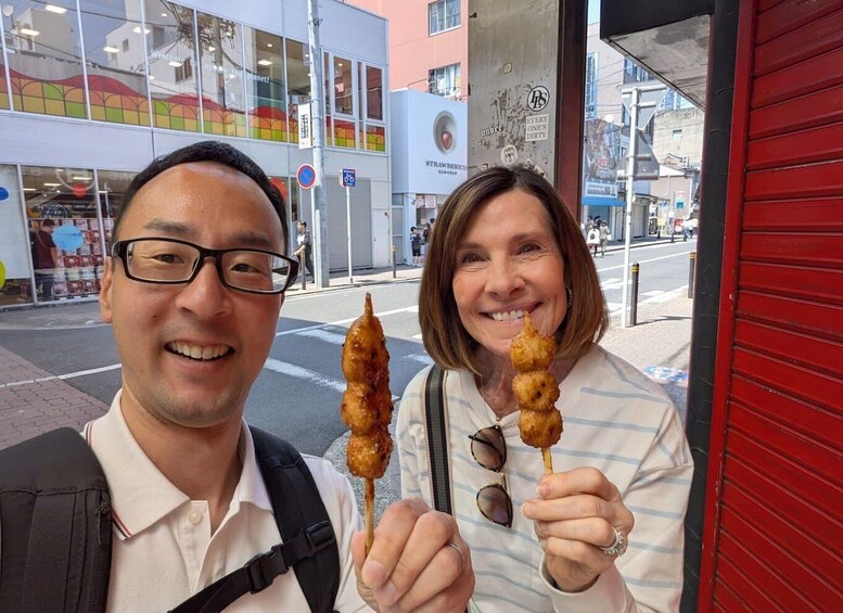 Two people holding skewers of food, with a building and a red door in the background. 2