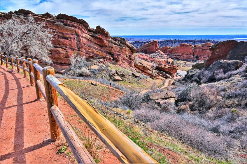 A red rock trail with wooden railings, sparse vegetation, and a distant view of the ocean. 1