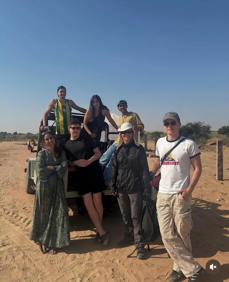 A group of people standing and sitting around a vehicle in a desert landscape. 2