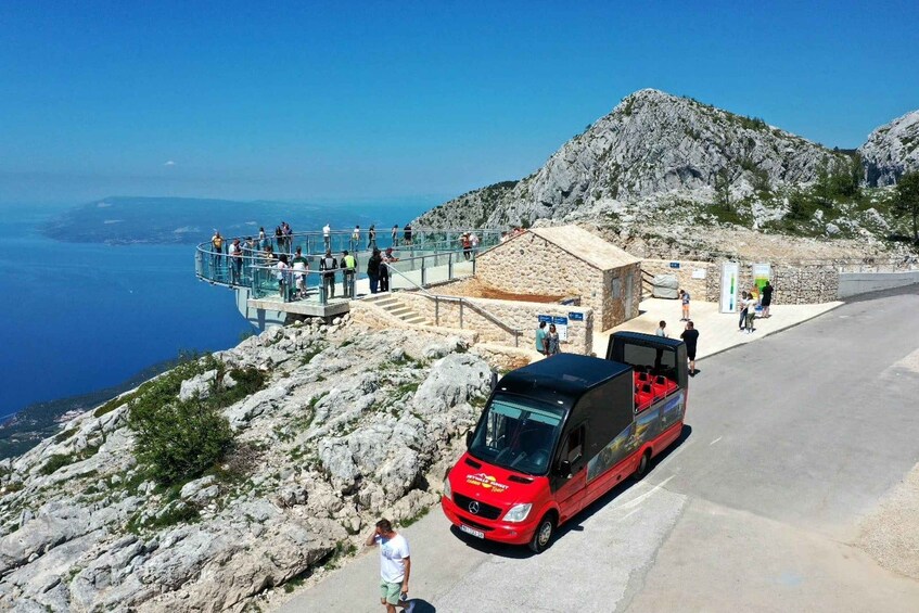 A red bus parked on a road with a rocky mountain and a clear blue sky in the background. 1