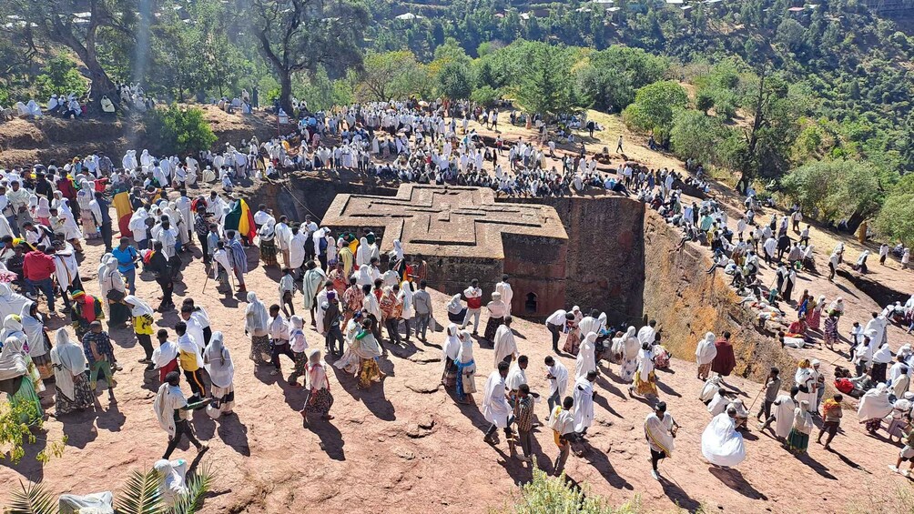 A large gathering of people around a central structure with a cross design, surrounded by trees and hills. 2