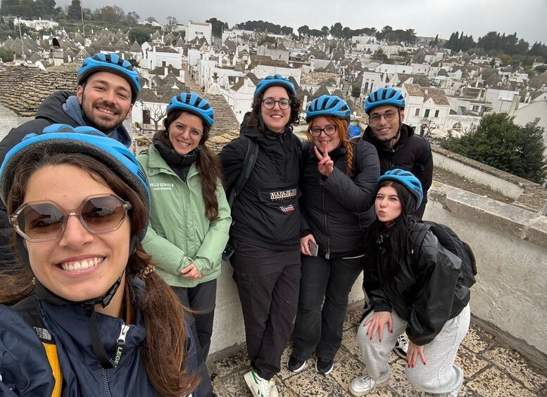 A group of people wearing helmets and jackets, standing on a stone wall overlooking a town with white buildings. 5