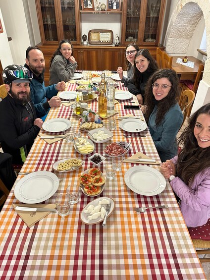 A group of people seated around a table with a checkered tablecloth, plates, glasses, and various food items. 3