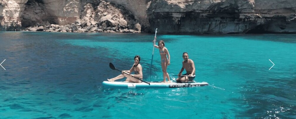 Three people on a paddleboard in clear turquoise water with rocky cliffs in the background. 3