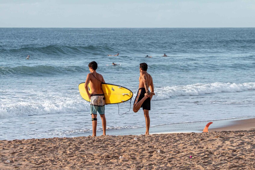 Two people on a beach with a yellow surfboard, ocean waves, and swimmers in the distance. 3
